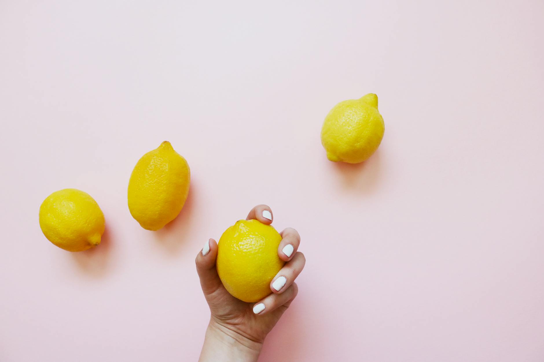 A hand holding a bright yellow lemon on a soft pink background
